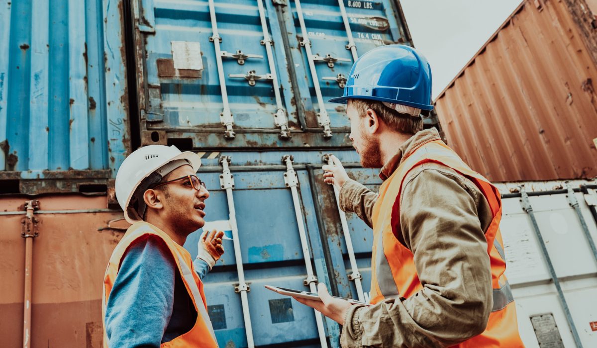 Two confident supervisors wearing reflective vests and hardhats discussing details of    loading ship with container storage units while gathered together in port